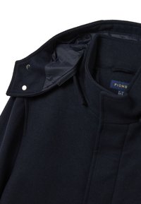 Cappotto in lana blu navy con colletto alto, chiusura con cerniera nascosta e bottoni automatici. Interno con fodera liscia a contrasto. Design minimalista.