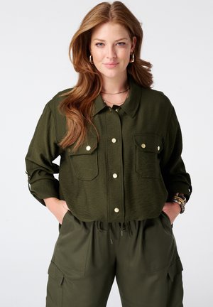 Vrouw met lang rood haar, die een olijfgroen overhemd met knopen en bijpassende cargobroek draagt, staat met handen in de zakken en glimlacht lichtjes.