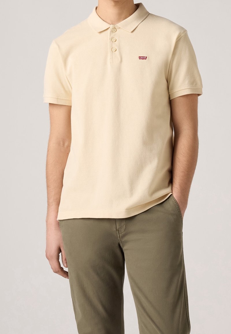Polo beige en tissu doux avec un col, comportant trois boutons et un petit logo rouge Levi's sur la poitrine. Porté avec un pantalon vert olive.