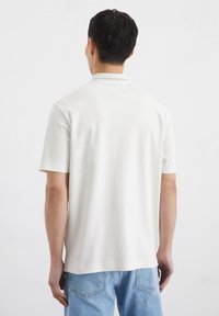 Hemd mit kurzen Ärmeln, aus weißer Baumwolle und mit einem klassischen Kragen und minimalistischen Design. Auf der Rückseite befindet sich ein kleines Etikett am Halsbereich. Kombiniert mit blauer Jeans.