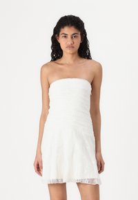 Robe blanche sans bretelles avec un tissu en dentelle superposée, jupe fluide et texture subtile, se terminant au-dessus du genou. Design de corsage lisse et ajusté.