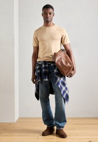 Camiseta beige de cuello redondo, camisa de cuadros azul atada a la cintura, jeans de mezclilla azules, bolso de cuero marrón y botas de tacón marrón sobre un suelo de madera.