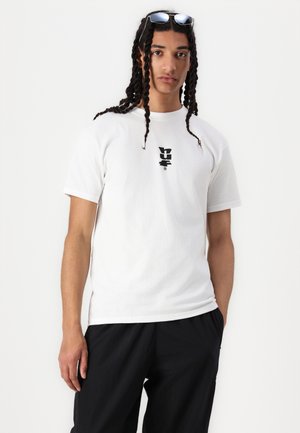 Hvid bomulds-t-shirt med et sort grafisk logo på brystet, rund hals og korte ærmer. Matchet med sorte løstsiddende bukser.