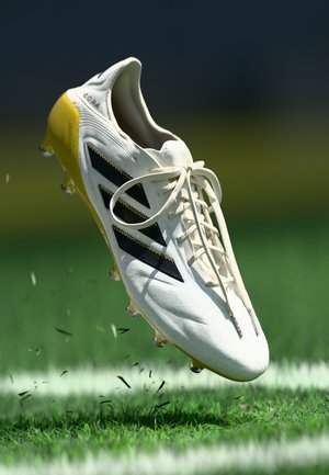 White soccer cleat with black stripes and yellow outsole, featuring a lace-up front and textured upper. Floating above green grass.