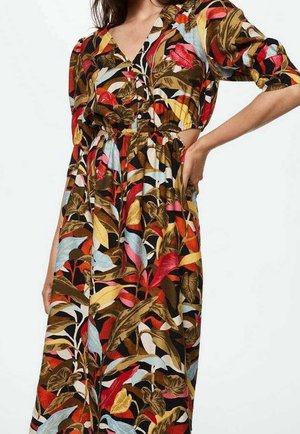 Frau trägt ein buntes, langärmliges Kleid mit einem lebhaften Blumen- und Blattmuster in Rot-, Gelb-, Blau-, Rosa- und Brauntönen.