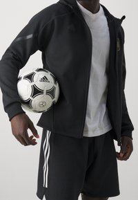 Homme en veste noire Adidas et short tenant un ballon de football Adidas blanc contre le côté, portant un T-shirt blanc sous la veste.