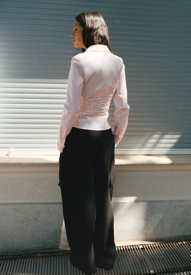 Femme aux longs cheveux foncés portant une veste ajustée rose clair et un pantalon noir ample, debout de profil contre un bâtiment avec des volets fermés.