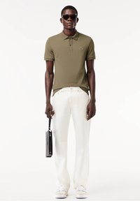 Homme portant un polo vert olive, un pantalon blanc, des baskets blanches et des lunettes de soleil noires tenant une pochette noire sur fond blanc.