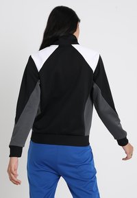 Svart och grå jacka med hög krage, med vita axelaccenter och dragkedjestängning. Texturerat material med ribbade ärmslut och nederkant.