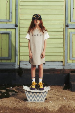 Grå klänning med vita ärmar och "MARNI" meddelande, gula och lila sneakers, stående på en mönstrad blå och vit plattform. Grön bakgrund.