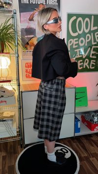 Svart kort jacka, rutiga knälånga kjol, grå solglasögon, vita strumpor och svarta skor. Minimalistisk inredning med affischer och växter.