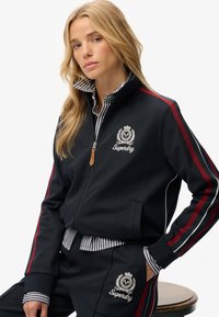 Marine-blauwe zip-up trainingsjack met rode en groene zijstrepen, voorzien van geborduurd logo en zakken, gecombineerd met een gestreept shirt met kraag.