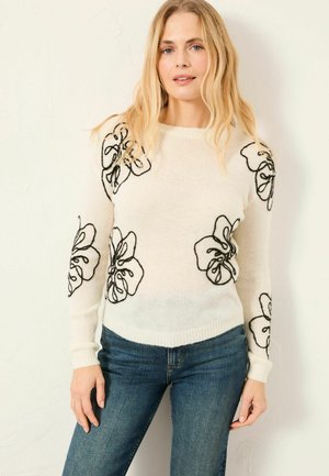 Valge kampsun musta lilleornamendiga varruka ja esiküljel, soonikkoes mansettide ja äärega. Kandatud koos siniste teksadega, sileda tekstuuriga.