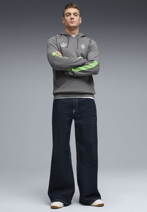 Noor mees seisab käed rüpes, kandes halli kapuutsiga dressipluusi roheliste triipudega, tumedaid laia jalaga teksapükse ja valgeid tossusid, halli ühevärvilise tausta ees.