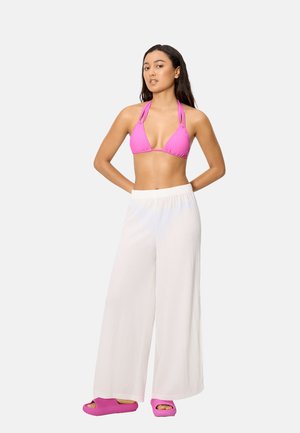 Mujer con top de bikini rosa, pantalones blancos de pierna ancha y sandalias rosas tipo slide, de pie con las manos detrás de la espalda sobre fondo blanco.