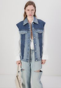 Denim vest with a patchwork design, featuring blue and white polka dots, large pockets, and silver snap buttons, worn over an oversized shirt.