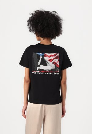 T-shirt en coton noir avec un imprimé graphique d'une femme devant un drapeau américain, avec "LOS ANGELES EST. 1981" en texte blanc en bas.