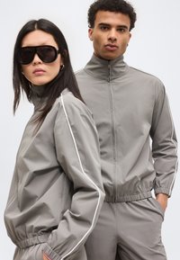 Kaks mudelit, kes kannavad ühesuguseid halli lukuga spordirõivaid valgete äärtega; üks prillidega, mõlemad seisavad ühetaustal heleda tausta ees.