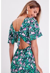 Vestito floreale verde con maniche a volant corte, design con schiena scoperta e dettaglio con fiocco. Presenta motivi di fiori rosa e bianchi sparsi in tutto il vestito.