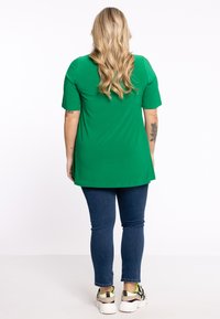 Grünes Kurzarmoberteil mit abgerundetem Saum, kombiniert mit dunkelblauen Slim-Fit-Jeans und weißen Sneakers mit schwarzen und neonfarbenen Akzenten.