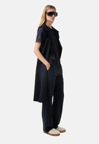 Marineblaues Outfit mit einem ärmellosen Weste, Satin-Oberteil, weiten Hosen und cremefarbenen Slippern, ergänzt durch übergroße dunkle Sonnenbrillen.