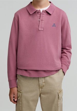 Kind trägt ein langärmeliges, staubrosa Poloshirt mit kleinem Totenkopf-Logo und beigefarbenen Cargohosen, steht mit einer Hand in der Tasche.