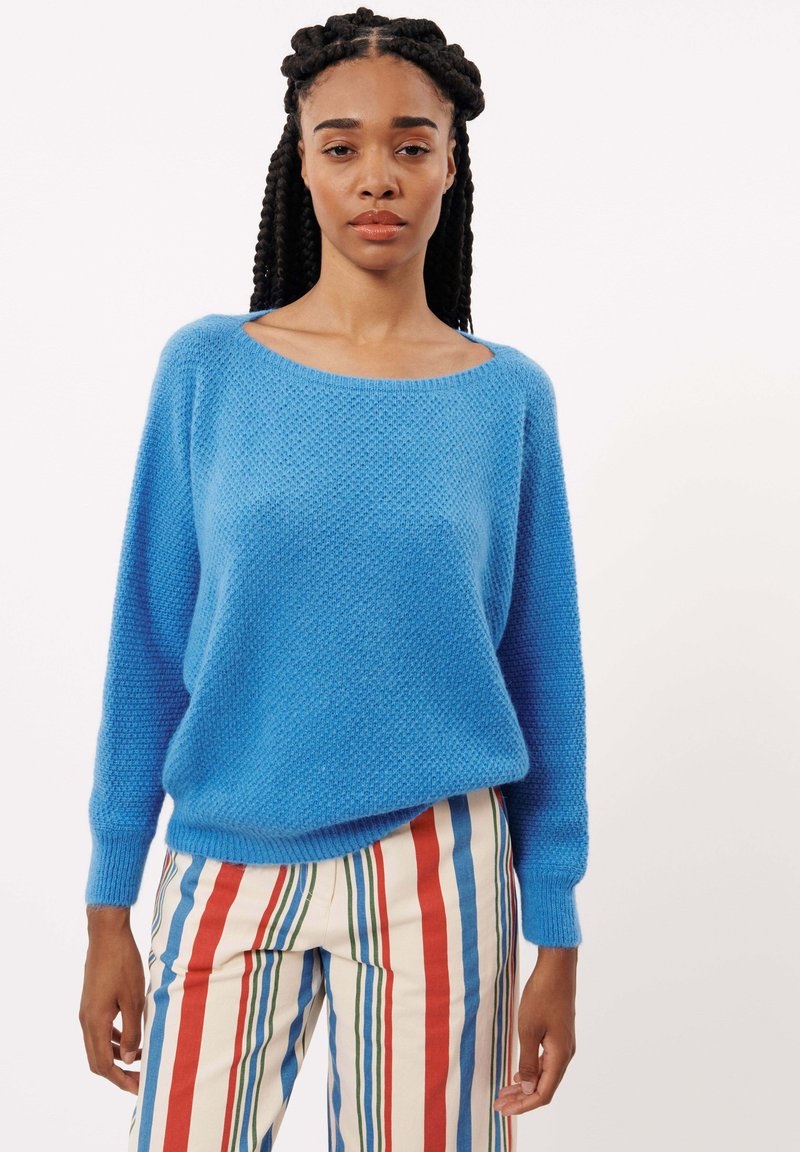 Pull en tricot bleu ciel avec un grand col et des manches longues ; associé à un pantalon crème à rayures comportant des rayures rouges, bleues et vertes.