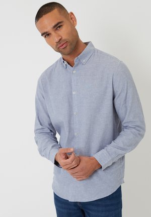 Man met kort haar, gekleed in een lichtblauw overhemd met knopen en lange mouwen, en een blauwe spijkerbroek, staand tegen een effen witte achtergrond.