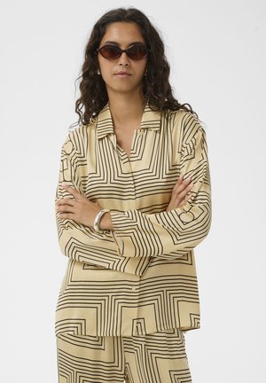 Pikkade lokkis juustega naine, kes kannab tumedaid päikeseprille, suure suurusega beeži särki ja mustade geomeetriliste triipudega pükse, hõbedast käevõru, käed ristis.