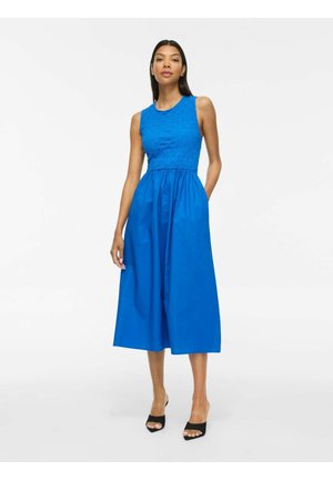 Femme en robe midi bleu vif sans manches avec haut texturé et jupe froncée, portant des sandales noires à talons ouvertes, debout sur fond blanc.