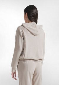 Sudadera beige con capucha, de ajuste holgado, cinturilla elástica y mangas largas. Textura suave, sin patrones visibles ni elementos metálicos.