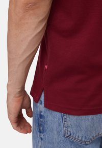 Mužova levá ruka a ruka má uvolněné u těla, má na sobě vínovou košili a světle modré džíny s viditelným prošíváním na zadních kapsách.