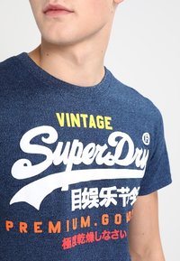 Mörkblå kortärmad t-shirt med rund hals. Har vit och orange tryckt text: "Vintage SuperDry" och "Premium.Good."