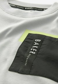 Šedé tričko s obdélníkovou černou kapsou, na které je bílý text "BAKER" a limetkově zelený čtvercový akcent. Bavlněný materiál, design s kulatým výstřihem.