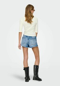Witte geribbelde top met driekwartmouwen, lichtblauwe denim shorts met afgeknotte pijpen en zwarte kniehoge laarzen met gespen, gezien van de achterkant.