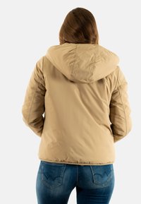 Giubbotto imbottito beige con cappuccio, texture trapuntata e orlo elastico. Abbinato a jeans in denim blu con cuciture decorative sulle tasche.