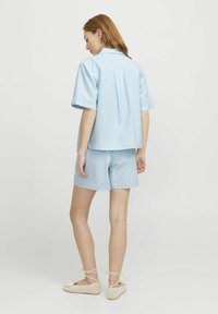 Camicia azzurra a righe con maniche corte e piega posteriore, abbinata a pantaloni corti coordinati. Completato con sandali intrecciati e accessori minimali.