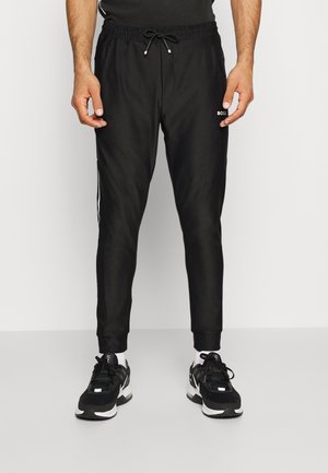 Hombre con joggers negros con cordón, logo BOSS en el muslo y zapatillas deportivas negras con detalles en blanco.