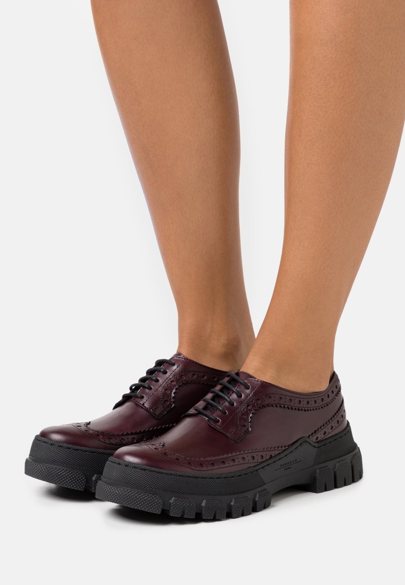 Chaussures brogues bordeaux foncé brillantes avec semelles épaisses en caoutchouc noir, portées sur jambes nues sur fond blanc.