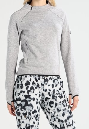 Grauer Fleecepullover mit hohem Kragen und Reißverschlusstaschen an den Seiten, kombiniert mit weißen Cargo-Hosen mit schwarzem Leopardenmuster.