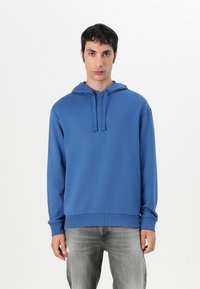 Modra flisasta hoodie s kapuco na vrvico, z rebrastimi manšetami in robom, ki ima majhen logotip na prsih, v kombinaciji s sivimi džinsom.
