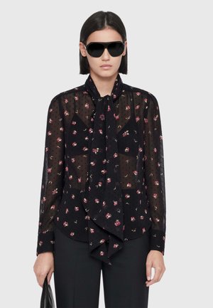 Woman wearing black sheer floral blouse with necktie, black pants, large black sunglasses, and holding a black handbag.