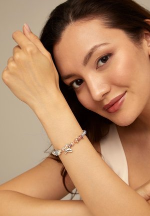 Mujer con maquillaje suave que lleva una pulsera de cuentas con amuletos de peces plateados, apoyando su brazo cerca del rostro y sonriendo suavemente.