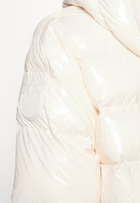 Puffjacka i glänsande elfenbensfärgad tyg med fastsydd huva och en upphöjd, rund logopatch på vänster arm. Har kviltad design.