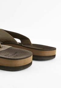 Brūni sintētiski flip-flopi ar teksturētu pēdu daļu, ar biezu, kontrastējošu brūnu starpzoli un tumšu ārējo zoli; minimālistisks dizains bez rakstiem.