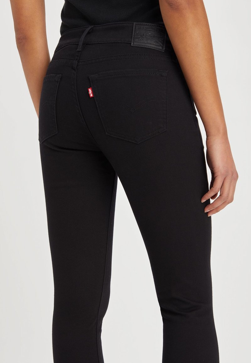 Schwarze Jeans aus Denim mit hoher Taille, ausgestattet mit zwei Gesäßtaschen und einem roten Logo-Tag. Die Textur ist glatt mit dezenten Nähdetails.