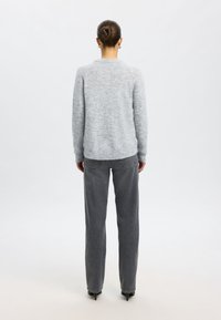 Femme avec les cheveux en chignon, portant un pull gris clair, un jean gris foncé à jambes droites et des chaussures à talons noires, debout de dos sur un fond blanc.