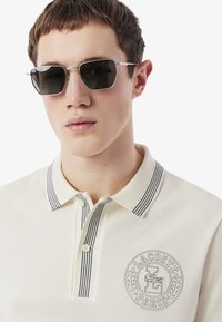 Világos bézs színű pólóing fekete csíkos gallérral és Lacoste logóval. A modellen sötét napszemüveg van ezüst részletekkel és minimalista kialakítással.