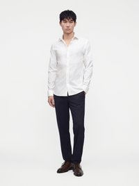 Uomo che indossa una camicia bianca a maniche lunghe con bottoni, pantaloni blu scuro e scarpe eleganti marroni, in piedi davanti a uno sfondo chiaro e uniforme.
