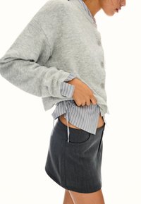 Kvinna som justerar en lager-på-lager-outfit med grå knappad cardigan, randig skjorta och mörkgrå kjol mot en vit bakgrund.